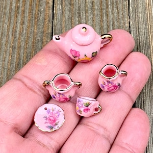 May include: Miniature pink porcelain tea set with gold trim, featuring a teapot, two creamers, a teacup, and a saucer. Each piece is decorated with floral patterns in pink, purple, and yellow. The set is designed for a dollhouse or miniature collection.