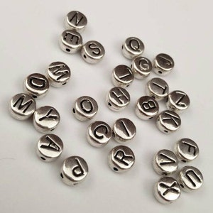 May include: A collection of silver metal alphabet beads with black lettering. The beads are arranged in a random pattern on a white surface.