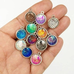 May include: Assortment of 12 iridescent mermaid scale charm pendants. Each pendant features a silver-toned metal frame with a round, iridescent glass cabochon. The cabochons feature a variety of colors, including blue, green, purple, pink, and orange.