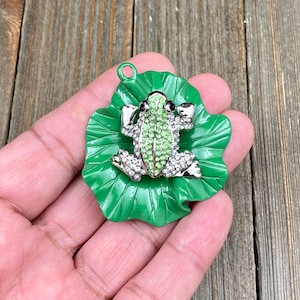 May include: A green and silver frog pendant with a green rhinestone body and clear rhinestone accents, resting on a green enameled lily pad. The pendant has a small loop at the top for attaching to a chain or other jewelry.