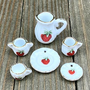 Puede incluir: Un juego de té de cerámica blanca en miniatura con ribetes dorados, con un diseño de fresa roja. El juego incluye una jarrita, tres tazas y dos platillos. Las piezas están dispuestas sobre una superficie de madera.
