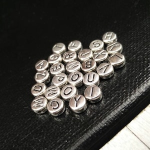 May include: A close-up of a pile of silver letter beads. The beads are round and have a smooth surface. Each bead has a different letter of the alphabet engraved on it.