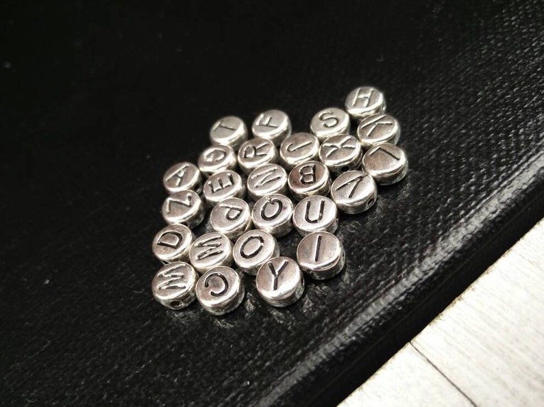 May include: A close-up of a pile of silver letter beads. The beads are round and have a smooth surface. Each bead has a different letter of the alphabet engraved on it.