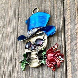 May include: A gold-toned skull charm with a blue top hat and a red rose in its mouth. The skull has red eyes and white teeth. The hat is decorated with rhinestones. The rose is also decorated with rhinestones.
