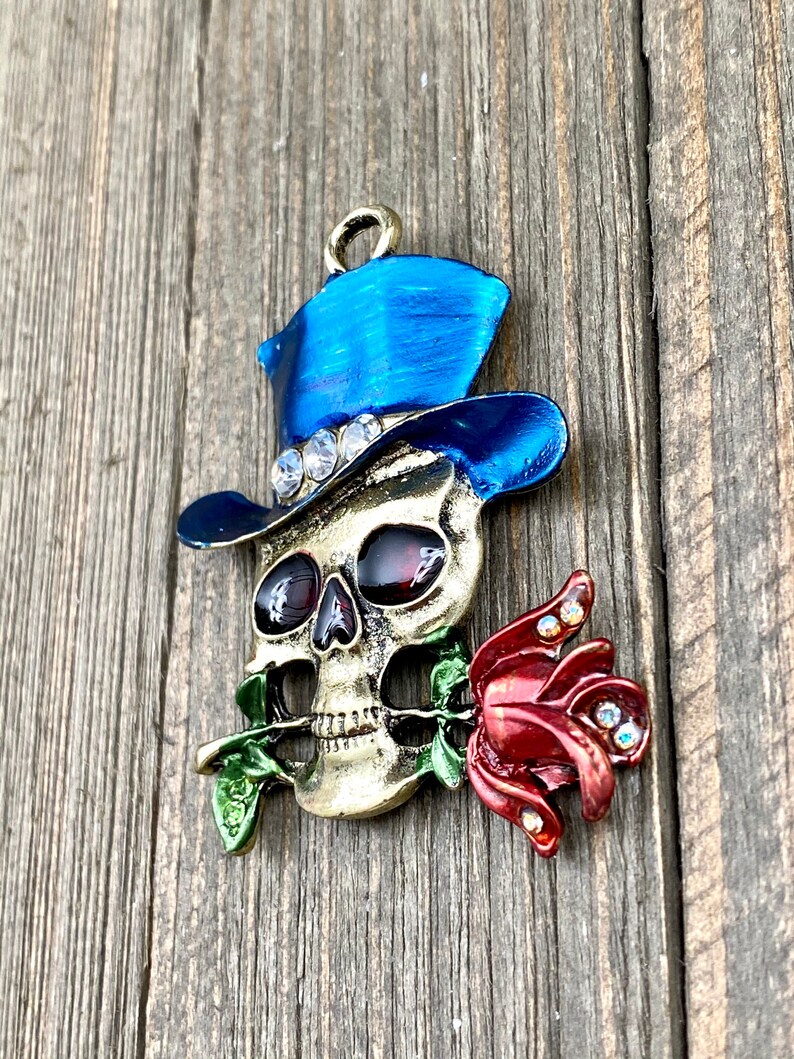 May include: A gold-toned skull charm with a blue top hat and a red rose in its mouth. The skull has red eyes and white teeth. The hat is decorated with rhinestones. The rose is also decorated with rhinestones.