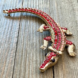 May include: A gold-toned alligator brooch encrusted with red and pink rhinestones. The brooch features a detailed design with a textured body and legs, and black eyes. The alligator's back is curved, and the brooch has a pin closure.