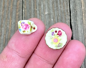 May include: Two miniature porcelain teacups with floral designs. The teacup on the left has a pink rose and green leaves. The teacup on the right is a saucer with a yellow flower and green leaves.