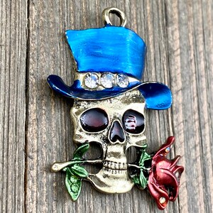 May include: A gold-toned skull charm with a blue top hat and a red rose. The hat has rhinestones and the skull has red eyes.
