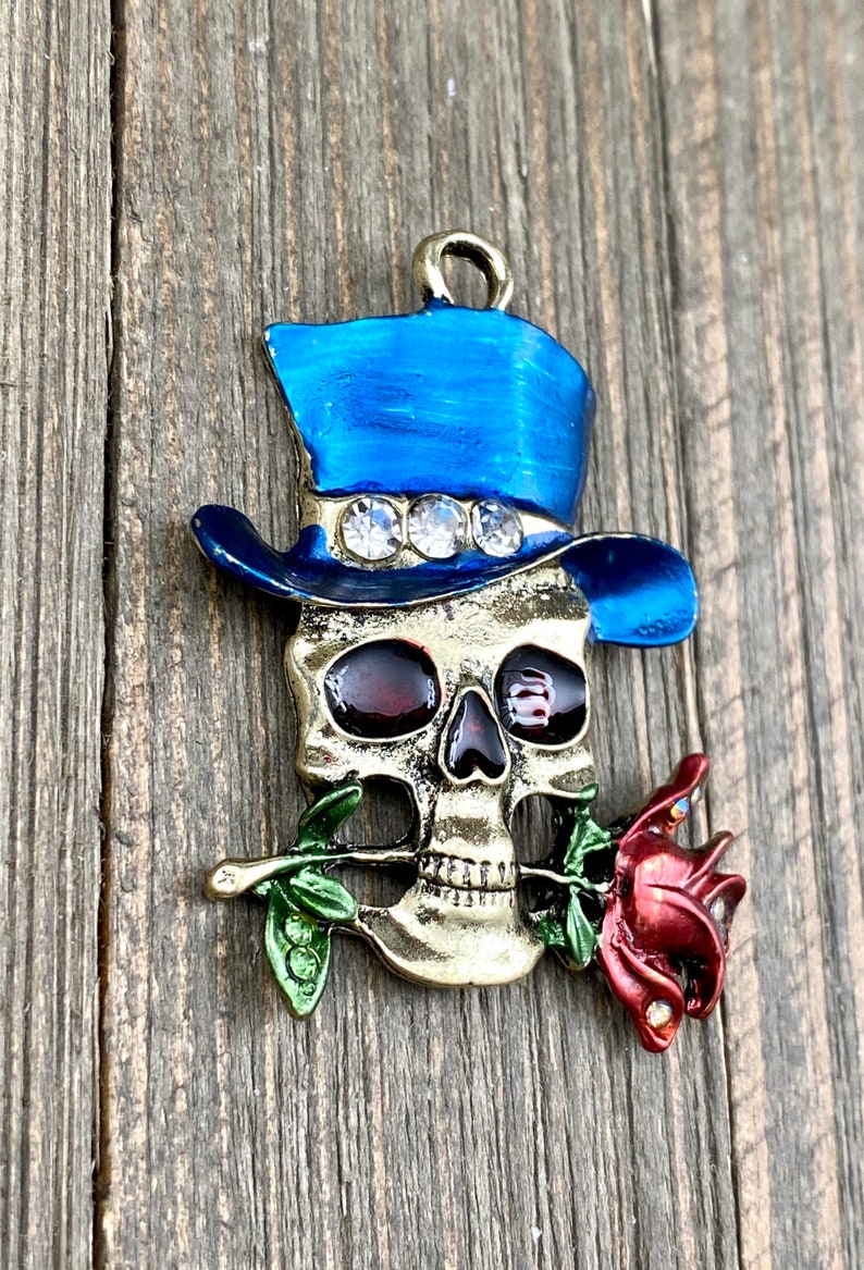 May include: A gold-toned skull charm with a blue top hat and a red rose. The hat has rhinestones and the skull has red eyes.