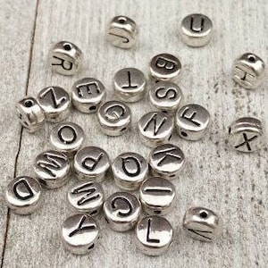 May include: A collection of silver metal alphabet letter beads. The beads are round and have a hole in the center for stringing. The letters are in uppercase and are all different.
