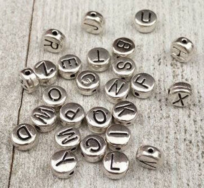 May include: A collection of silver metal alphabet letter beads. The beads are round and have a hole in the center for stringing. The letters are in uppercase and are all different.