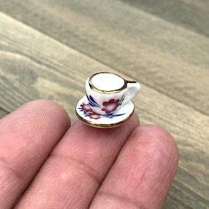 May include: A miniature teacup and saucer set with a floral design. The cup and saucer are white with a gold rim and feature pink, blue, and white flowers.