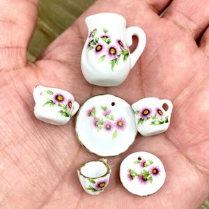 Puede incluir: Un juego de té en miniatura con un diseño floral. El juego incluye una jarra, dos tazas, dos platillos y un plato. El diseño floral presenta flores rosas y hojas verdes sobre un fondo blanco.