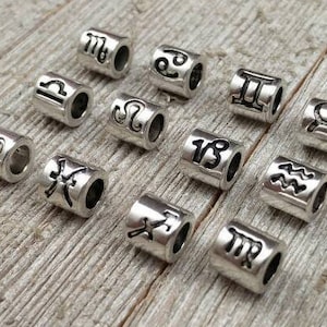 May include: Twelve silver metal beads with different zodiac symbols engraved on them. The beads are arranged in a semi-circle on a wooden surface.