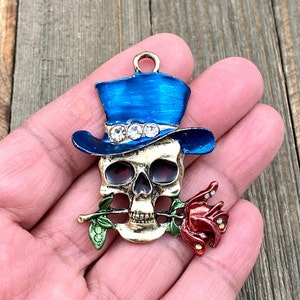 May include: A gold-tone skull charm with a blue top hat and a red rose. The skull has rhinestones on the hat and the rose has green leaves.