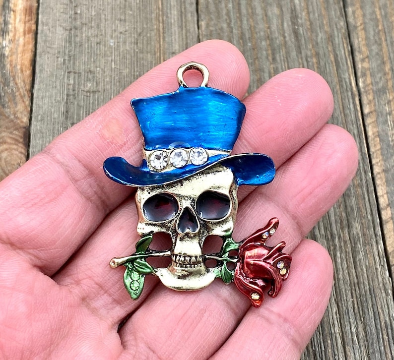 May include: A gold-tone skull charm with a blue top hat and a red rose. The skull has rhinestones on the hat and the rose has green leaves.