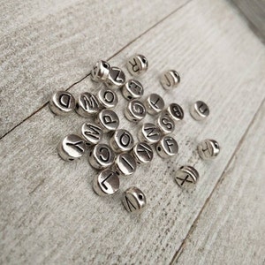 May include: A collection of silver letter beads with black lettering. The beads are scattered on a light wood surface.