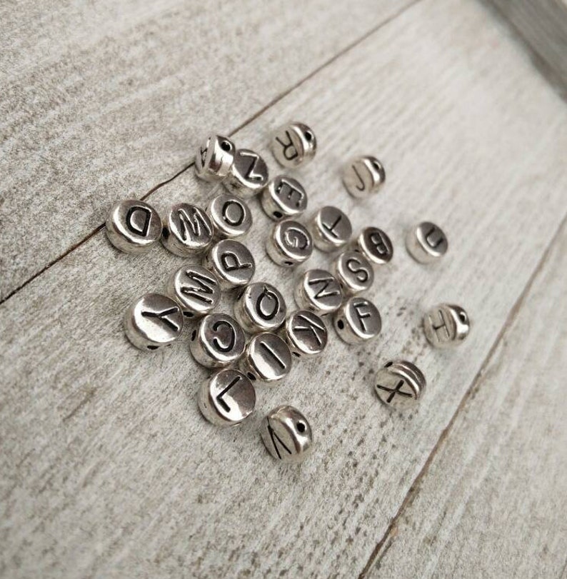 May include: A collection of silver letter beads with black lettering. The beads are scattered on a light wood surface.