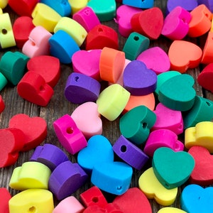 Puede incluir: Una colección de cuentas en forma de corazón de colores, en tonos de rojo, rosa, amarillo, verde, azul, morado y naranja. Cada cuenta tiene un agujero para ensartar. Las cuentas están dispersas, creando una exhibición vibrante y juguetona.