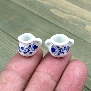 Puede incluir: Dos pequeños jarros de cerámica blanca con diseños florales azules. Los jarros tienen un asa en un lado y un pico en el otro.