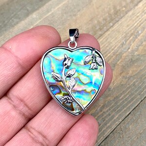 Peut inclure: Un pendentif en forme de cœur argenté avec un délicat motif floral. Le pendentif est fait de nacre d'ormeau aux reflets bleus, verts et dorés.