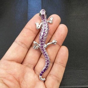 May include: A silver lizard brooch with purple enamel detailing. The lizard is posed with its legs outstretched and its tail curled.