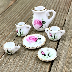 Puede incluir: Un juego de té de porcelana en miniatura con base blanca, que presenta diseños de rosas rosas y adornos dorados. El juego incluye una crema, azucarero, tazas y platillos. Las piezas están dispuestas sobre una superficie de madera.