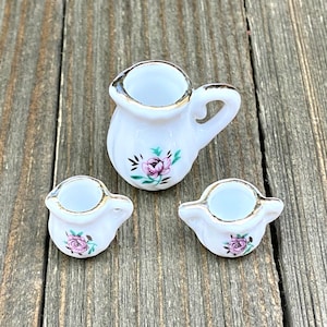 Puede incluir: Un juego de té de porcelana blanca con un diseño floral. El juego incluye una jarra y dos tazas.