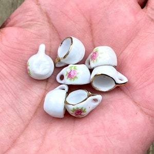 Może przedstawiać: Zestaw sześciu miniaturowych filiżanek do herbaty z porcelany z kwiatowymi wzorami. Filiżanki są białe z różowymi i zielonymi wzorami kwiatowymi i złotymi akcentami.