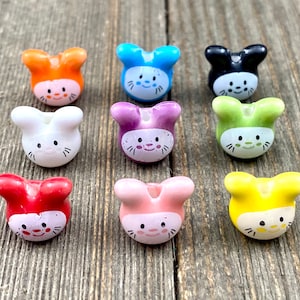 Puede incluir: Una colección de nueve cuentas de cerámica con forma de cara de conejo de colores. Cada cuenta presenta un color diferente, incluyendo naranja, azul, negro, blanco, morado, verde, rojo, rosa y amarillo. Las caras de los conejos tienen ojos y bigotes negros, y una nariz y mejillas rosas.