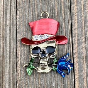 May include: A gold-toned skull charm with a red top hat and a blue rose. The skull has black eyes and a gold-toned nose and mouth. The rose has green leaves and is held in the skull's mouth.