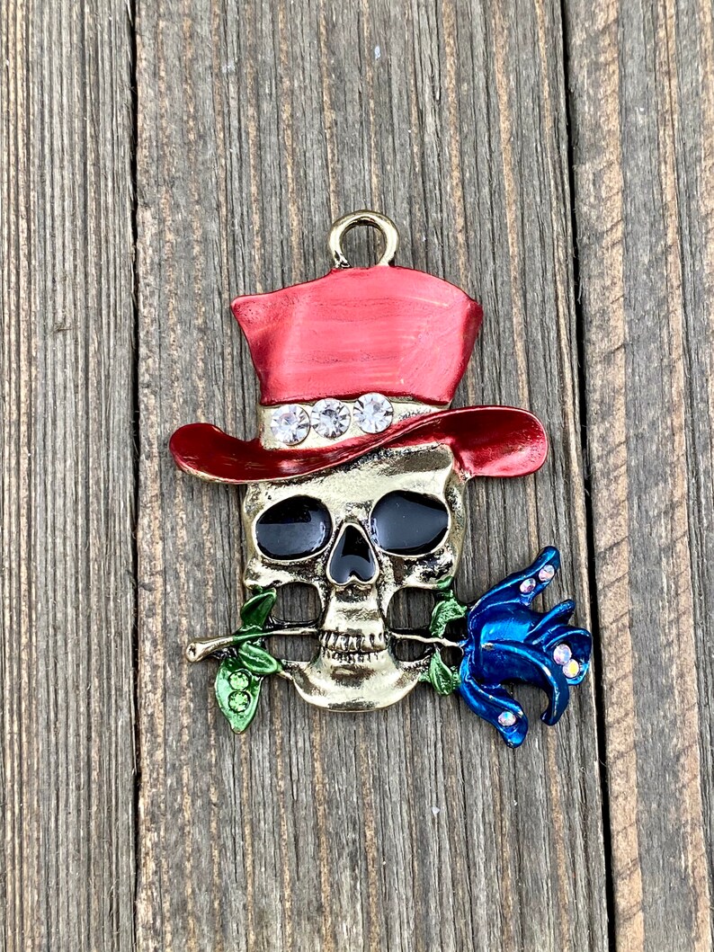 May include: A gold-toned skull charm with a red top hat and a blue rose. The skull has black eyes and a gold-toned nose and mouth. The rose has green leaves and is held in the skull's mouth.