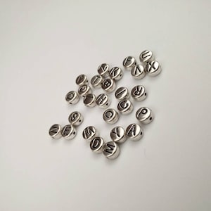 May include: A collection of silver letter beads with black lettering. The beads are scattered on a white surface.