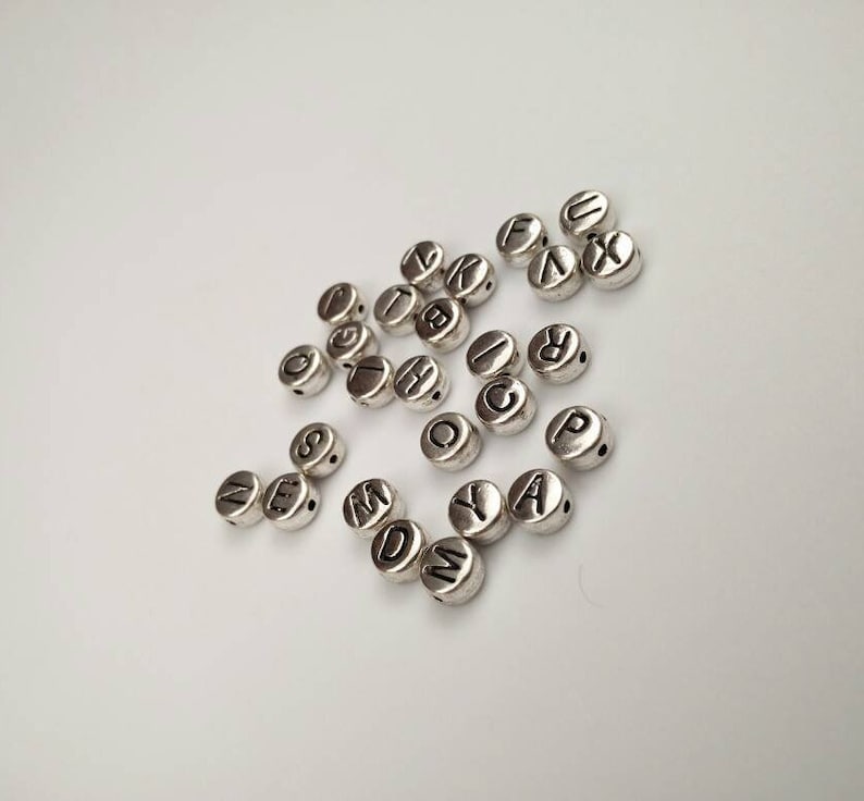 May include: A collection of silver letter beads with black lettering. The beads are scattered on a white surface.