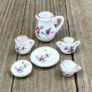 Puede incluir: Un juego de té en miniatura con una tetera blanca, tres tazas, un platillo y un plato. El juego está decorado con rosas rosas y hojas verdes.
