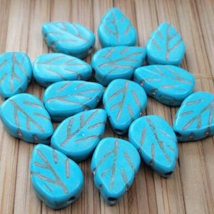 May include: A collection of turquoise blue glass beads shaped like leaves with a silver leaf design.