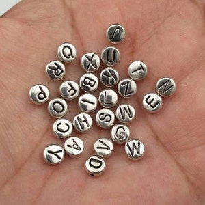 May include: A collection of silver-toned metal alphabet letter beads. Each bead is round and features a single letter of the alphabet in black.