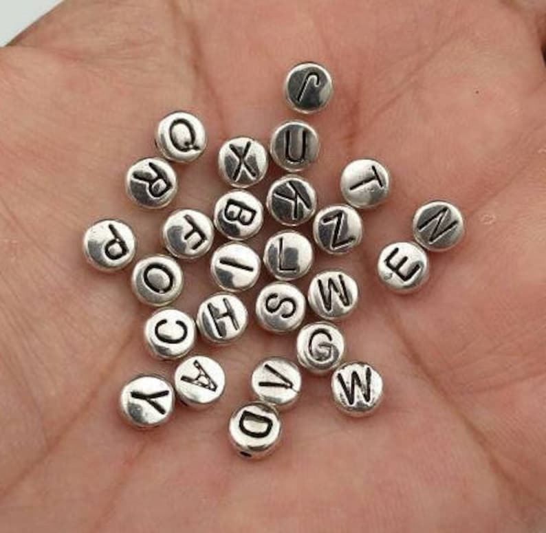 May include: A collection of silver-toned metal alphabet letter beads. Each bead is round and features a single letter of the alphabet in black.