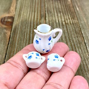 Puede incluir: Un juego de té en miniatura con una jarra blanca y dos tazas. El juego está decorado con flores azules y tiene un borde dorado.