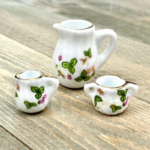 Puede incluir: Un juego de té en miniatura con una jarra blanca y dos tazas. El juego está decorado con un patrón floral de hojas verdes y flores rosas.