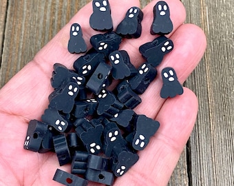 May include: A pile of black ghost-shaped polymer clay beads with white eyes. The beads have a hole through the top for stringing.