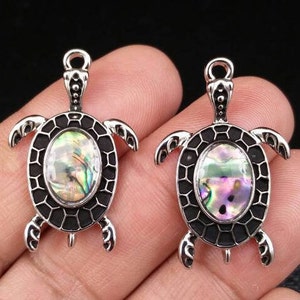 May include: Two silver-toned metal turtle pendants featuring an oval-shaped iridescent abalone shell inlay at the center of the shell. The turtle design includes a black recessed pattern on the shell and a small loop at the top for attachment.