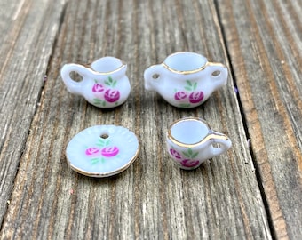 Puede incluir: Un juego de té en miniatura con una tetera blanca, una taza de té blanca y un platillo blanco. El juego está decorado con rosas rosas y hojas verdes.