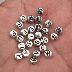 May include: Silver letter beads with black lettering. The beads are round and have a smooth surface. The letters are all uppercase and are evenly spaced on the beads.