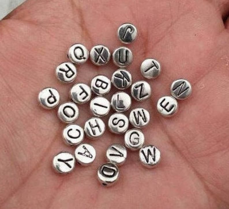 May include: Silver letter beads with black lettering. The beads are round and have a smooth surface. The letters are all uppercase and are evenly spaced on the beads.