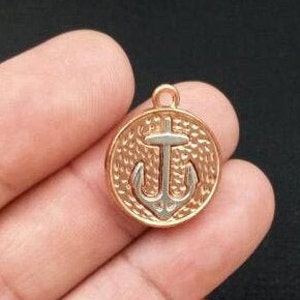 May include: A gold-toned metal charm with a silver anchor design. The charm is round and has a textured surface. It has a loop at the top for attaching to a necklace or bracelet.
