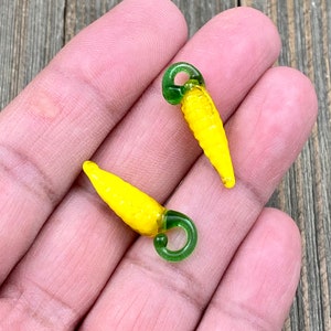 Puede incluir: Dos abalorios de vidrio amarillo en forma de pimiento con tapas verdes. Los abalorios tienen un bucle en la parte superior para sujetarlos a joyas u otras manualidades.