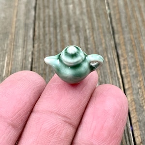 May include: A miniature ceramic teapot with a green and white swirl design. The teapot has a small spout and handle.
