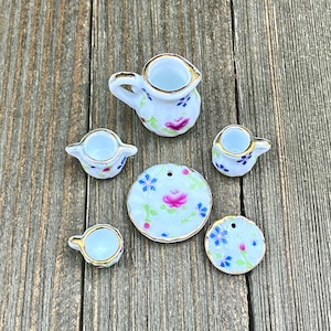 Puede incluir: Un juego de té en miniatura con una tetera blanca, dos tazas, dos platillos y un diseño floral. El juego está hecho de porcelana y tiene detalles dorados.