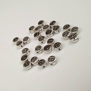 May include: Silver letter beads with black lettering. The beads are round and have a hole in the center. The letters are A through Z.